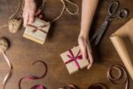 Hands tie pink ribbon around small kraft gift boxes on a wooden table, with scissors, twine, and wrapping paper nearby on the surface.