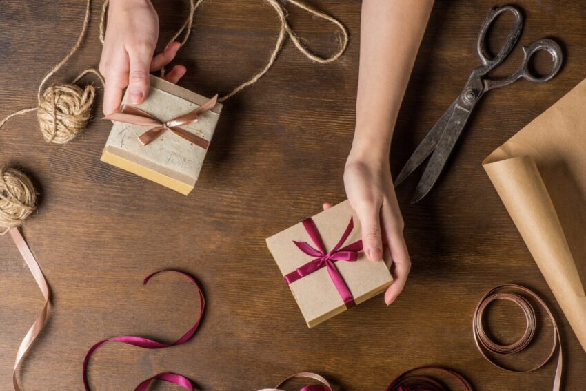 Hands tie pink ribbon around small kraft gift boxes on a wooden table, with scissors, twine, and wrapping paper nearby on the surface.