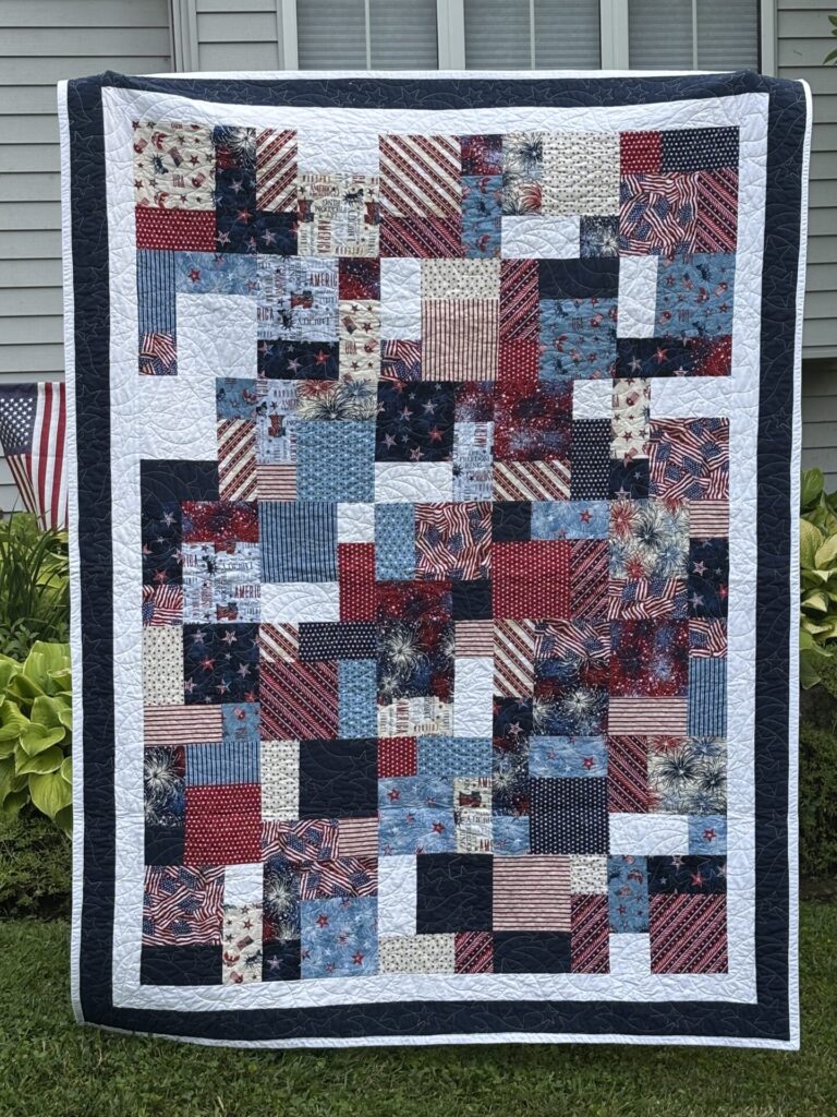 Patriotic patchwork quilt with red, white, and blue squares and starry border draped outside a house.