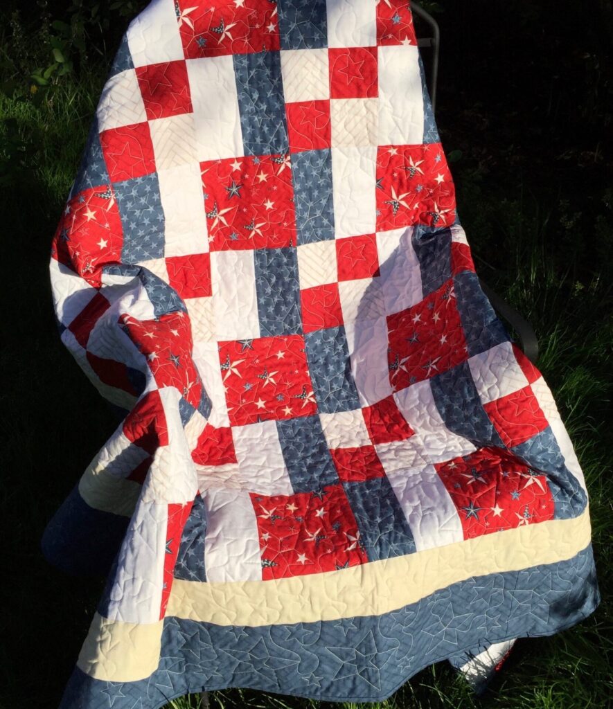 Patriotic quilt with red, white, and blue squares and star patterns draped over a chair outdoors.
