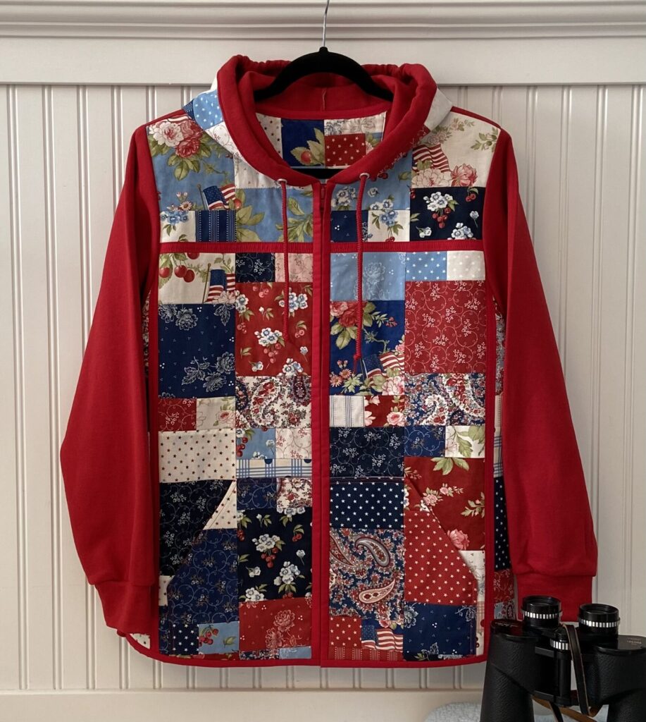 Red hoodie with a patchwork front featuring blue, red, and floral squares on a hanger. Detects a patriotic, quilt-like design.
