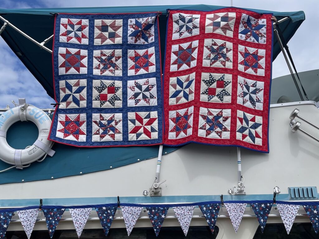 Two red, white, and blue patriotic quilts hanging over a boat railing, featuring star-block patterns on blue and red frames.