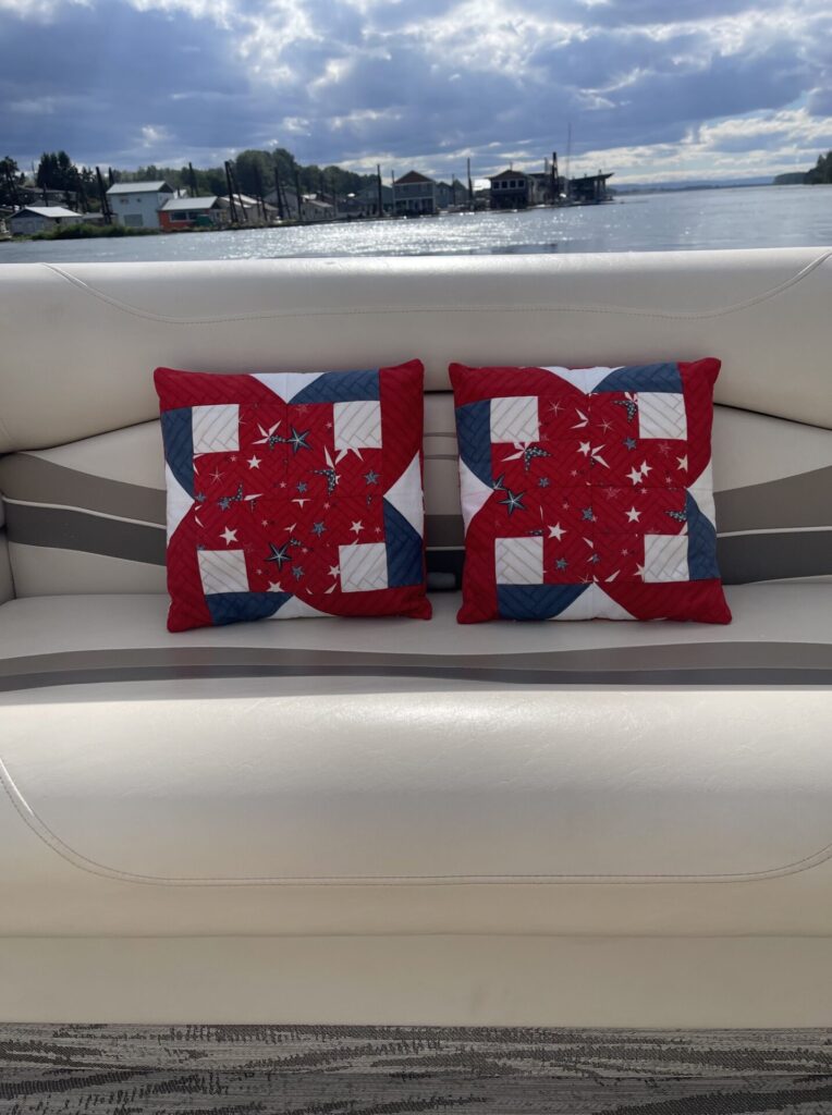 Two red patriotic quilt-pattern pillows sit on a white boat seat with a water view and shoreline houses in the distance.