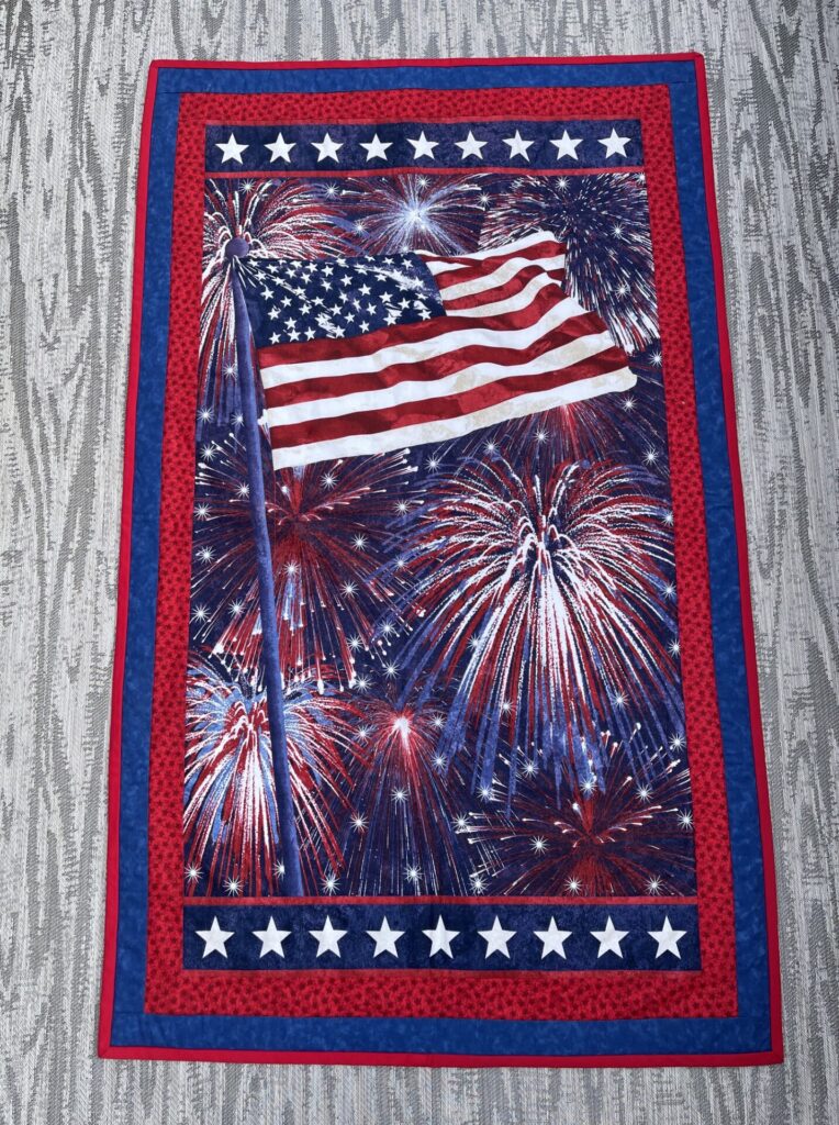 Patriotic quilt with a waving American flag over fireworks, framed by red and blue star-pattern borders.
