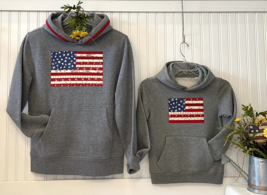 Two gray hoodies with red, white, and blue American flag patches hanging on a white beadboard wall. Floral accents peek from the top of the left hoodie.