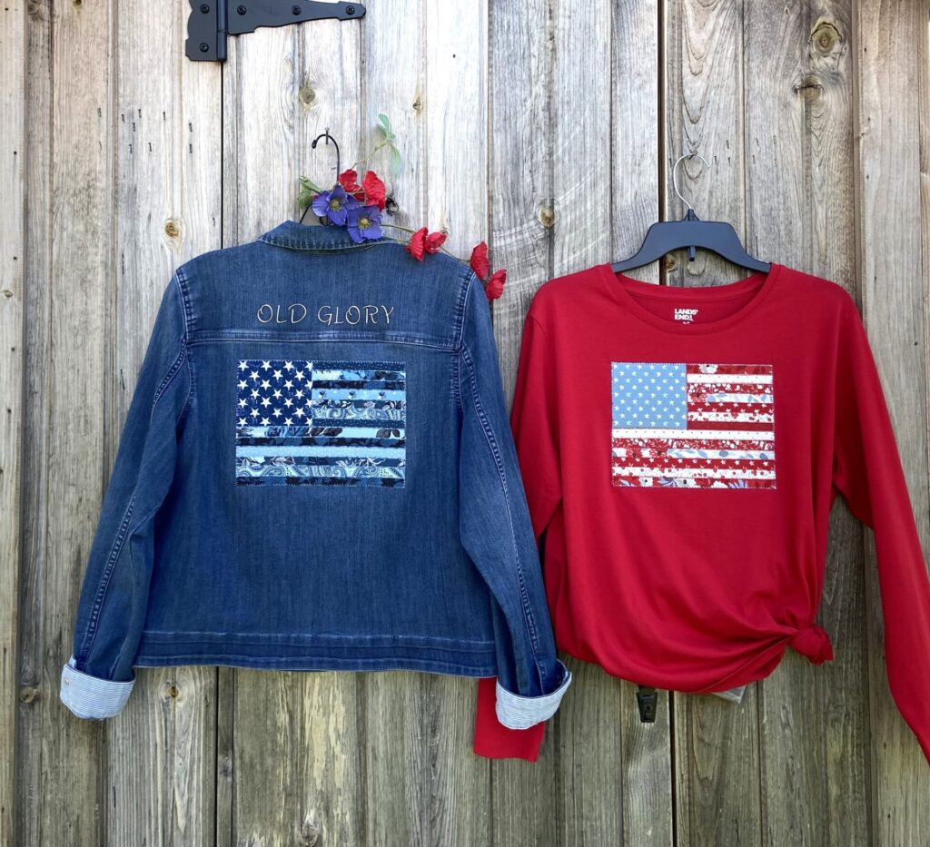 Patriotic clothing display on a weathered fence: a denim jacket with 'Old Glory' and an American flag patch beside a red shirt with a flag graphic on hangers.