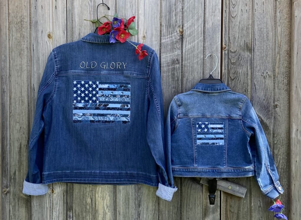 Two blue denim jackets hanging on a weathered wooden fence; one jacket features an embroidered 'OLD GLORY' and an American flag patch, with a colorful flower garland draped at the hanger edge.
