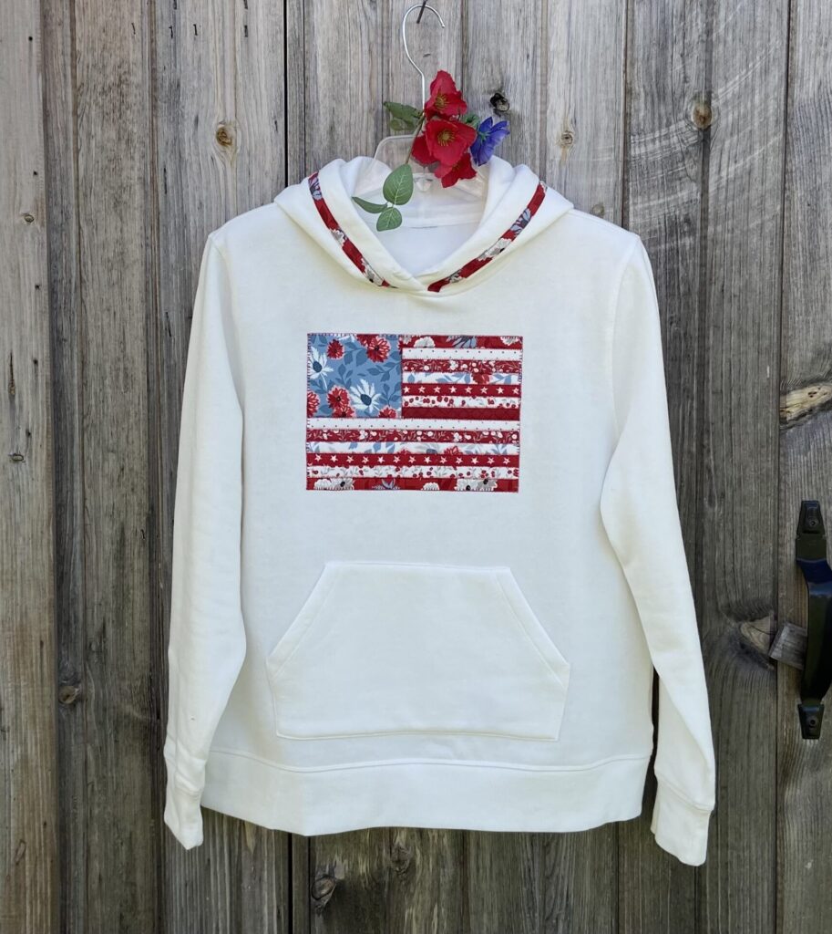 White hoodie with a patchwork American-flag motif on the chest, hung on a hanger with red flowers and greenery against a weathered wooden fence backdrop.