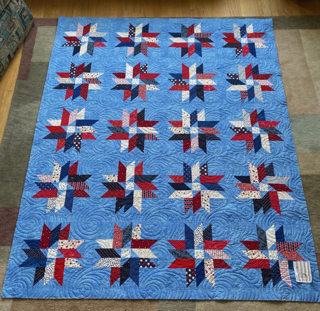 Patriotic quilt on the floor with red, white, and blue star blocks on a light blue swirled background.