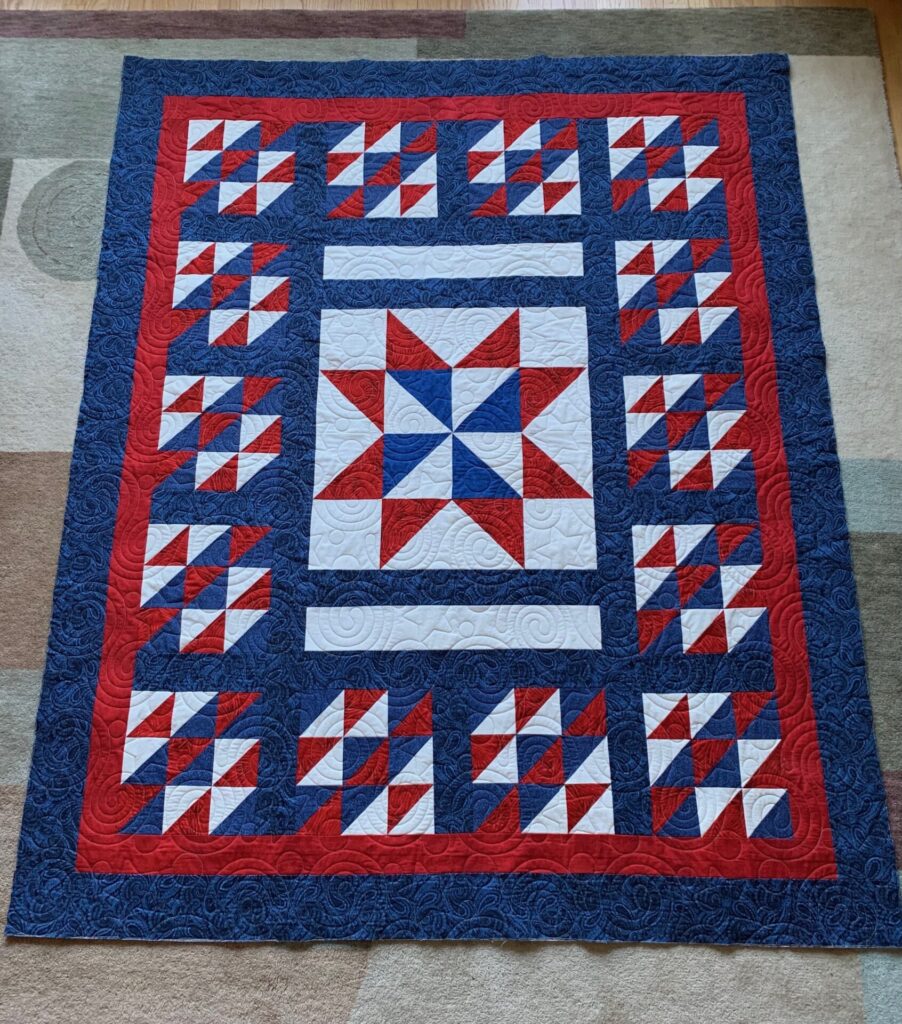 Patriotic quilt in red, white, and blue with star and pinwheel blocks and a swirled blue border on a red inner border.