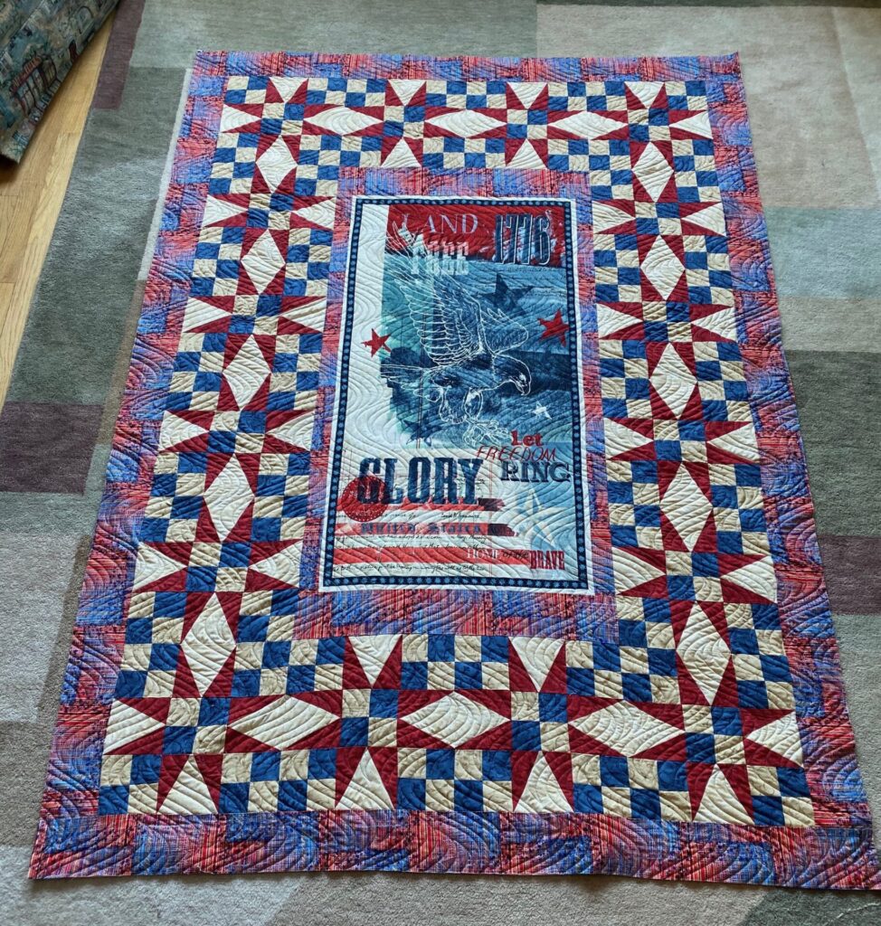 Patriotic quilt laid on a carpet, featuring red, white, and blue starburst blocks and a central panel with an eagle and the words 'Let freedom ring'.