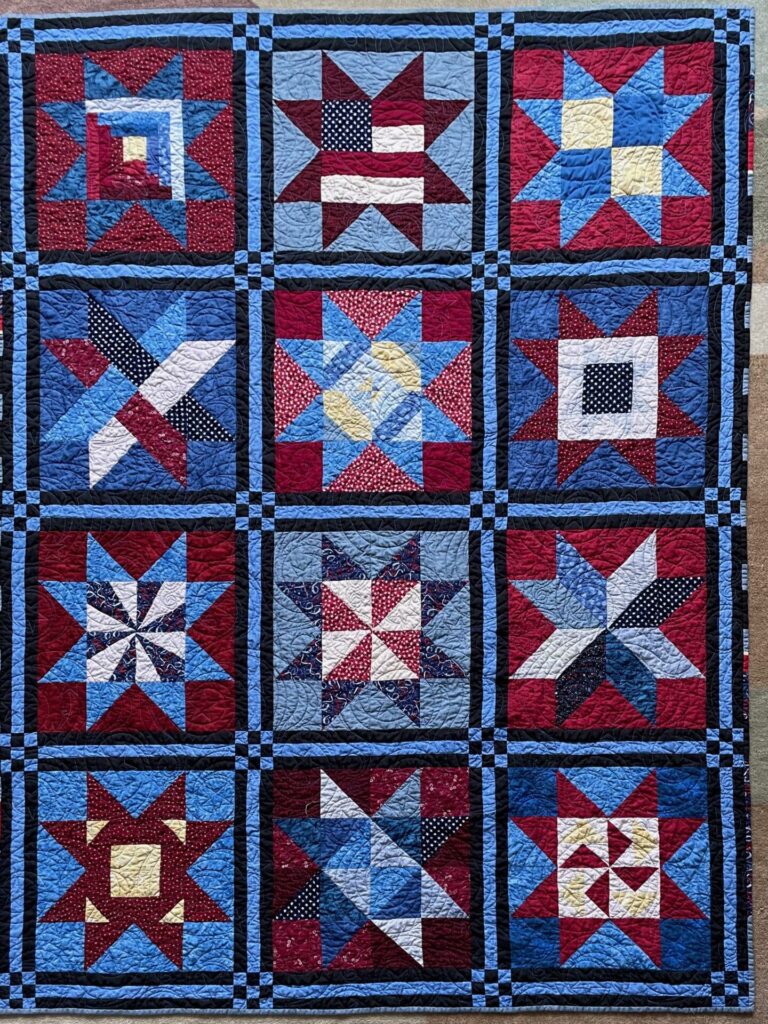 Patchwork quilt with nine star blocks in red, white, and blue, bordered by blue and black sashes (patriotic theme).