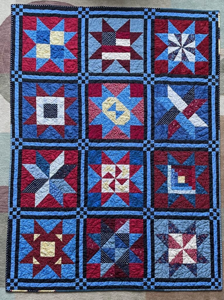Large quilt made of nine star blocks in red, white, and blue with dark blue borders and light inner squares.