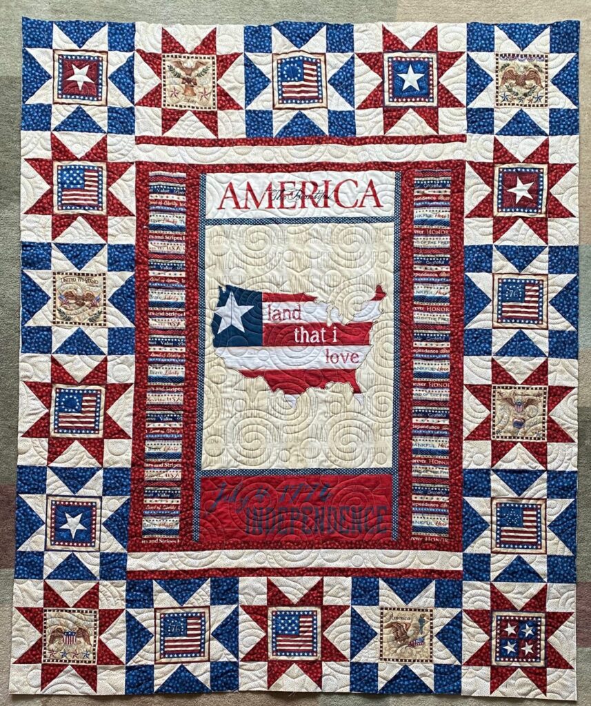 Patriotic quilt with red, white, and blue star blocks framing a central panel that reads AMERICA with map motifs and eagle squares.