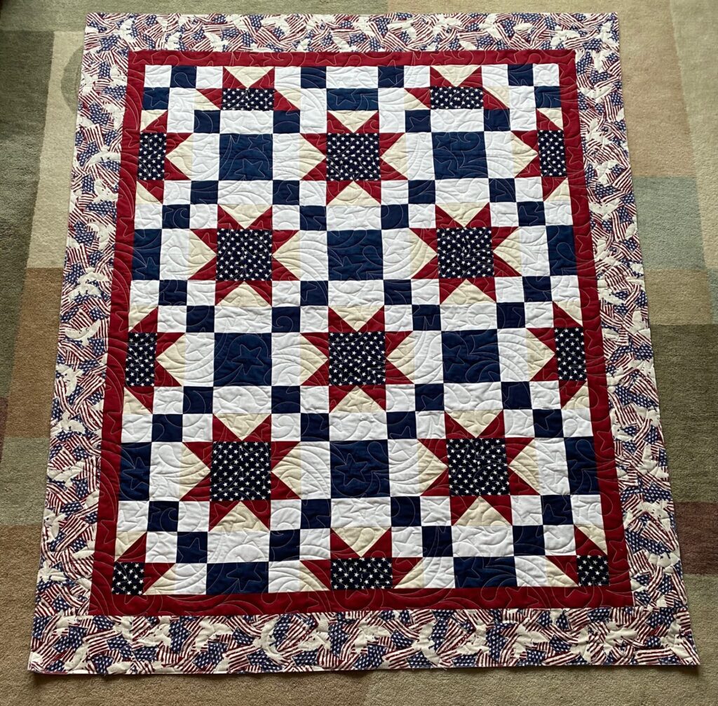 Patriotic quilt with red, white, and blue star and flag blocks and a flag-pattern border on a beige carpet.