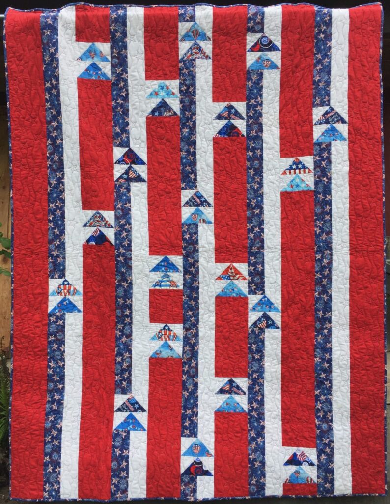 Patriotic quilt hanging vertically with red and white stripes, blue starry border strips, and tree-shaped blocks printed with USA-themed fabrics.