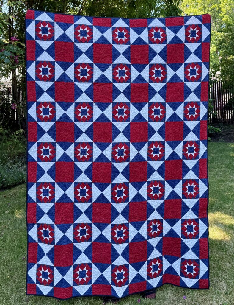 Red, white, and blue patriotic quilt with star-circle blocks on a checkerboard grid, hanging outdoors.