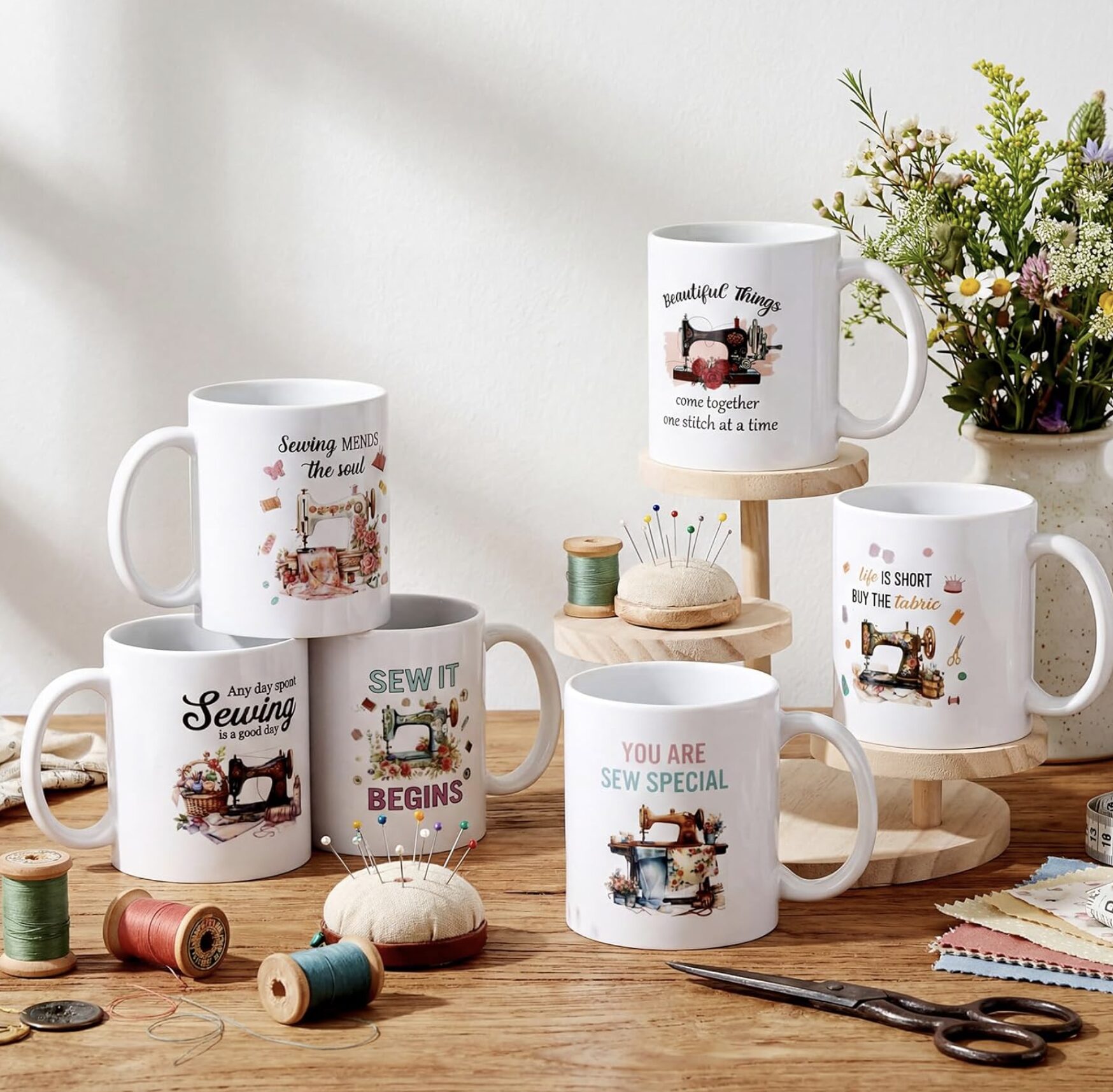 White mugs with sewing-themed quotes on a wooden display, surrounded by spools, pins, scissors and a bouquet of flowers.
