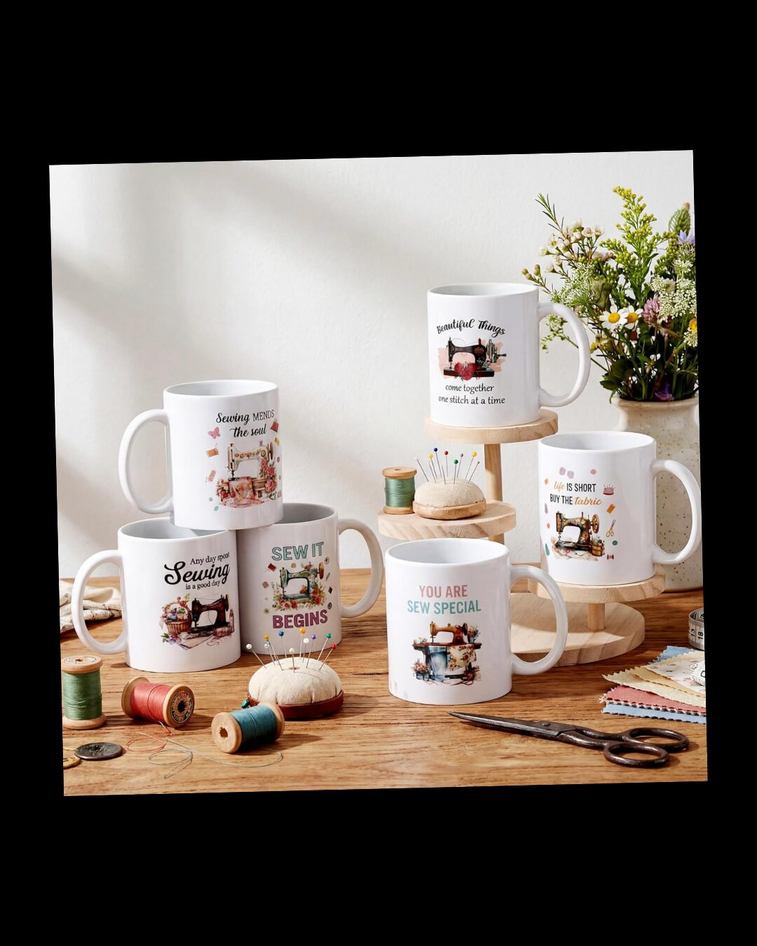 White mugs with sewing-themed designs and quotes on a wooden table, surrounded by thread spools, pins, scissors, and a bouquet of flowers.