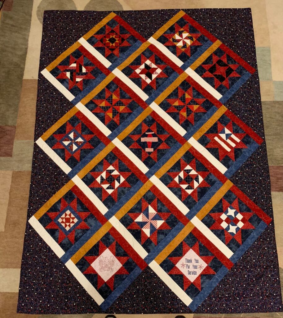 Quilt with diagonal diamond blocks in red, blue, white and gold sashing, bordered by a speckled dark frame; a small label in the bottom corner reads 'Thank you for your service'.