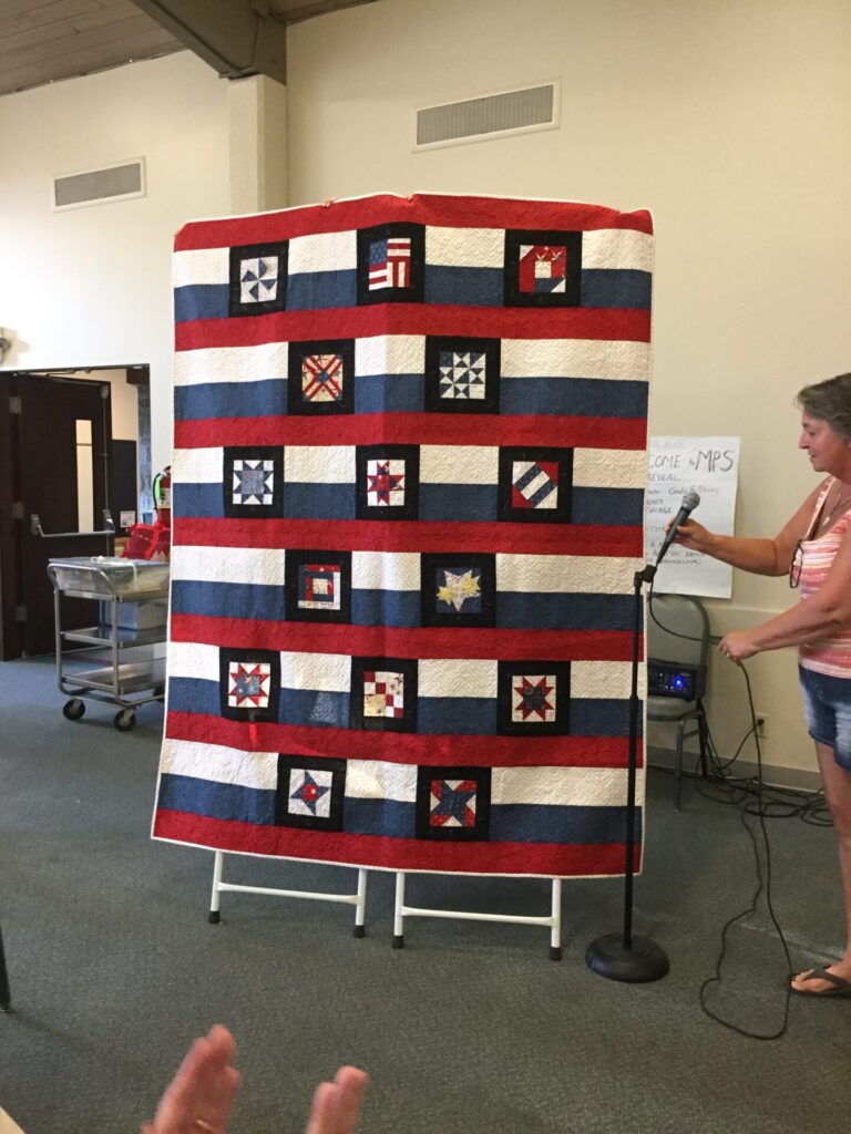 Red, white, and blue striped quilt with black-framed patchwork squares displayed on a stand in an indoor setting, with a presenter holding a microphone to the right.