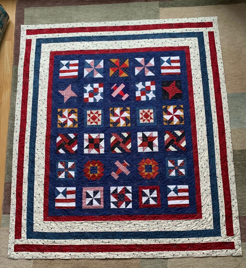 Patriotic quilt laid on the floor with blue, red, and white borders and multiple star- and flag-themed blocks.
