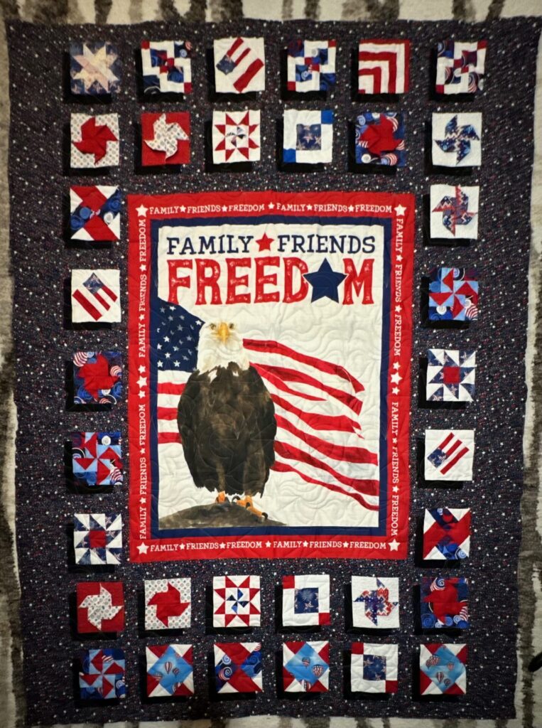 Patriotic quilt featuring a central panel with an eagle, stars and stripes, and the words FAMILY FRIENDS FREEDOM surrounded by blocks of red, white, and blue fabric.
