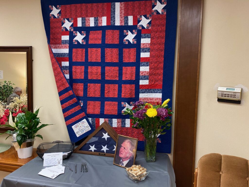 Patriotic quilt draped on a wall with blue, red, and white blocks and star accents, beside flowers and framed portraits.